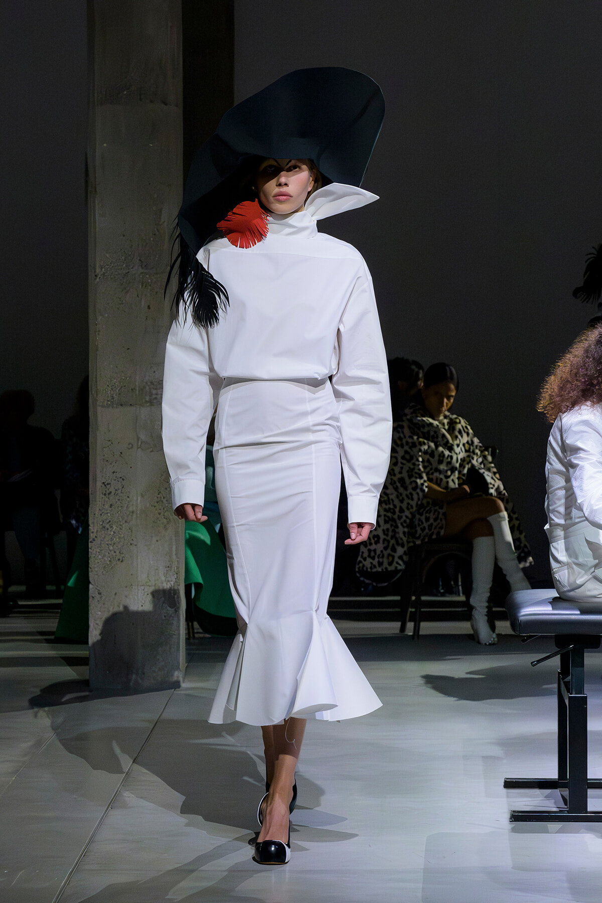 Model in a white high-neck outfit with an oversized black hat and a red feather accessory walking the runway.