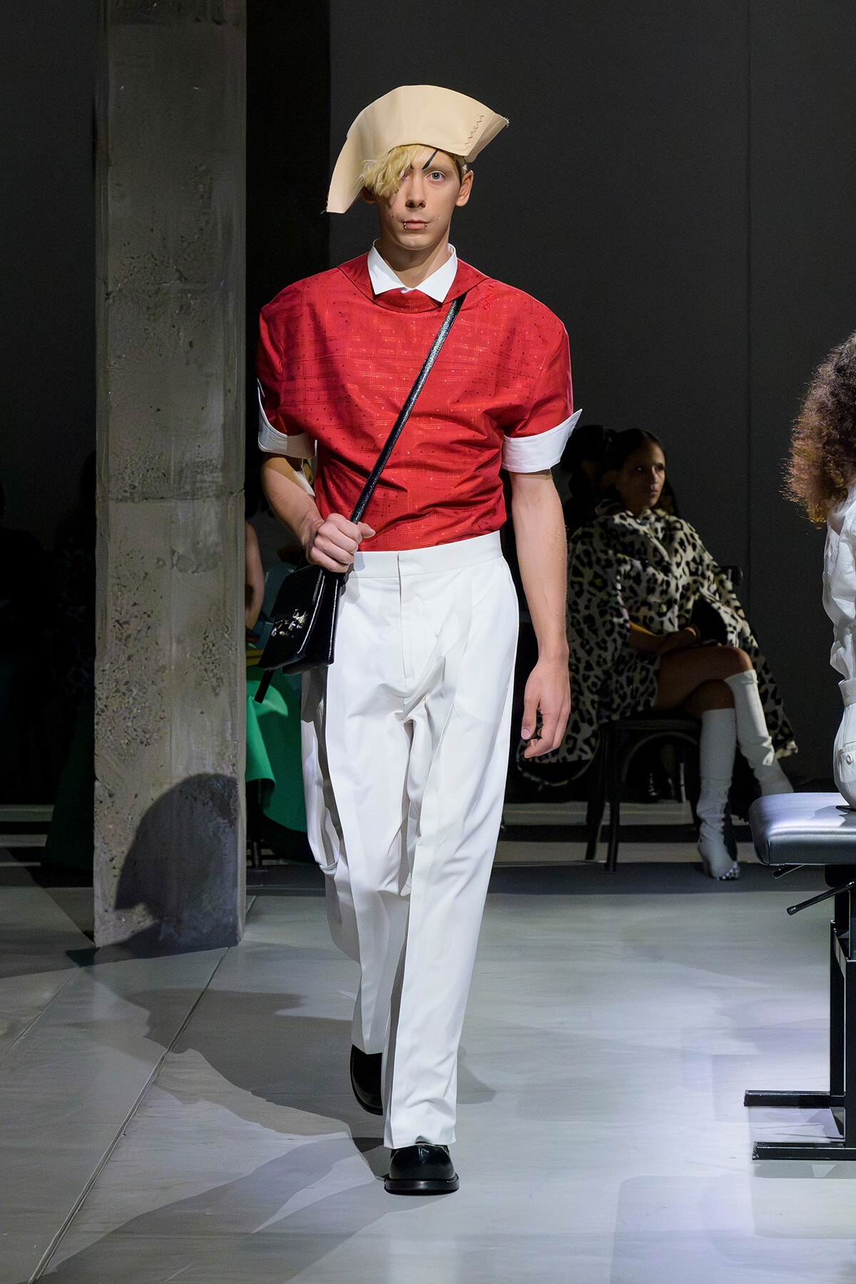 Male model walks a runway in a red short-sleeve shirt, white trousers, and a pale beige sculptural hat with a black crossbody bag.