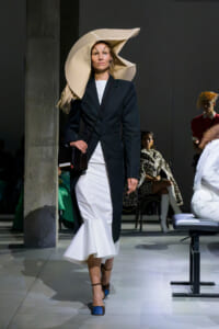 Model on the runway in a navy blazer over a white mermaid dress, with an oversized beige sculptural hat, black crossbody bag, and blue heels.