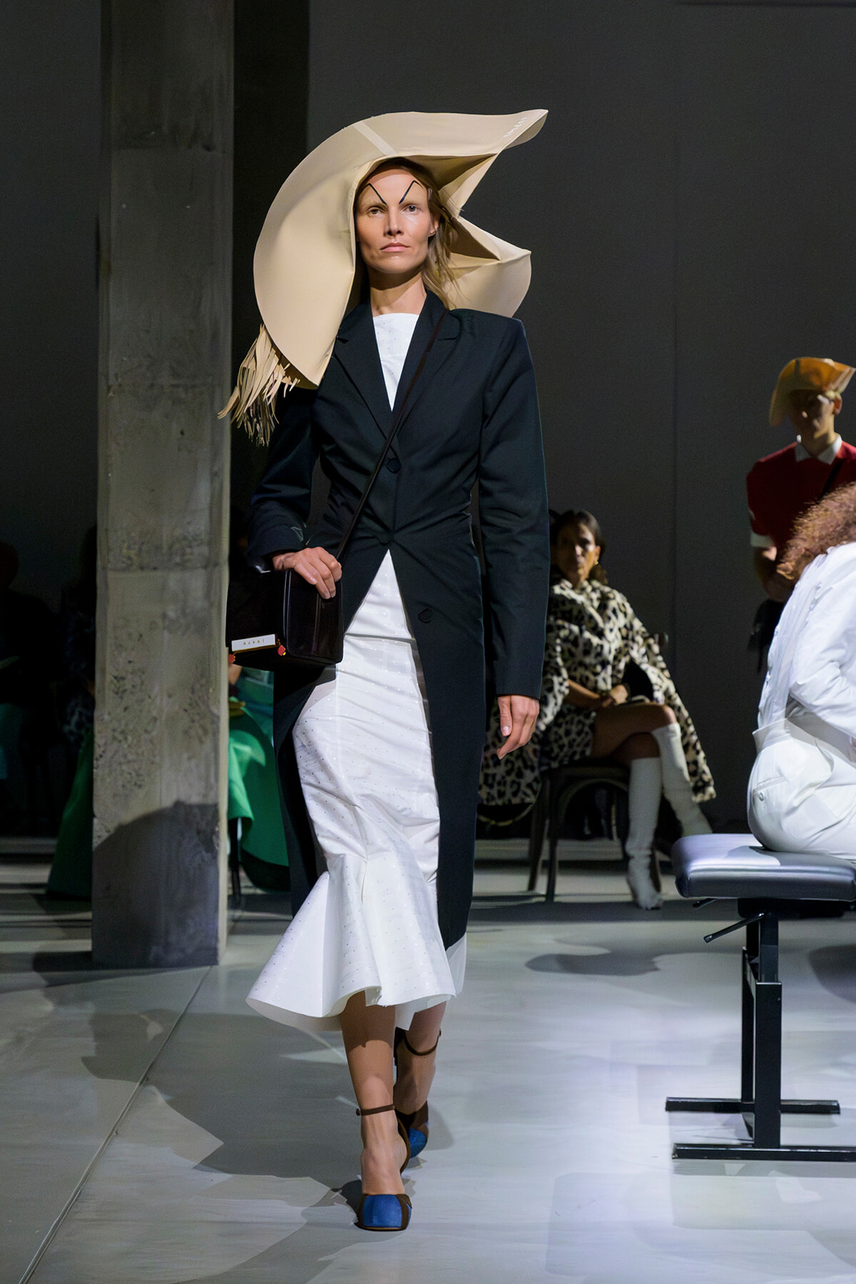 Model on the runway in a navy blazer over a white mermaid dress, with an oversized beige sculptural hat, black crossbody bag, and blue heels.