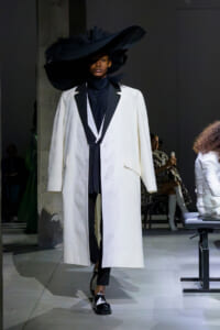 Runway model wearing a long white coat with black lapels over a black turtleneck, finished with black-and-white shoes and a dramatic oversized black hat.