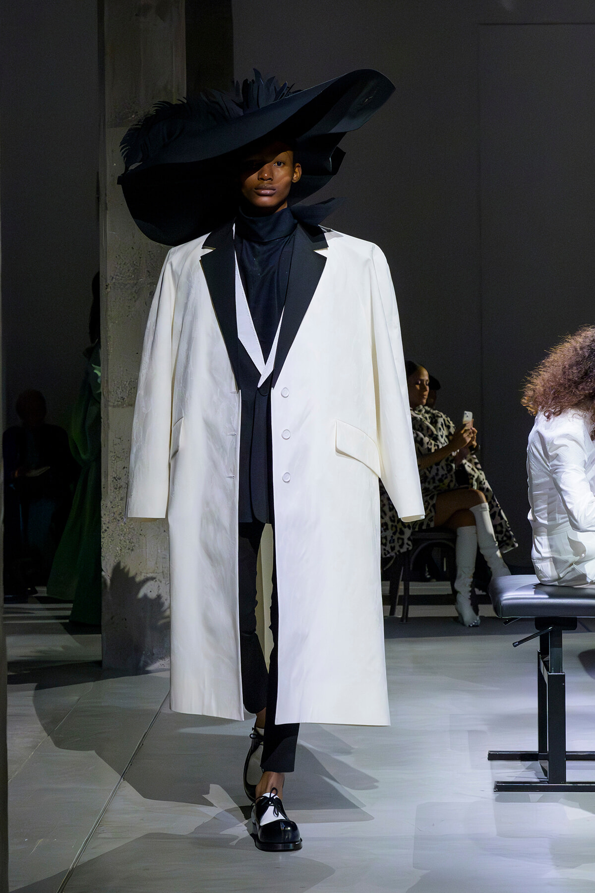 Runway model wearing a long white coat with black lapels over a black turtleneck, finished with black-and-white shoes and a dramatic oversized black hat.