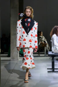 Model on runway wearing a white rose-print suit with black lapels and a dark scarf, walking the catwalk.