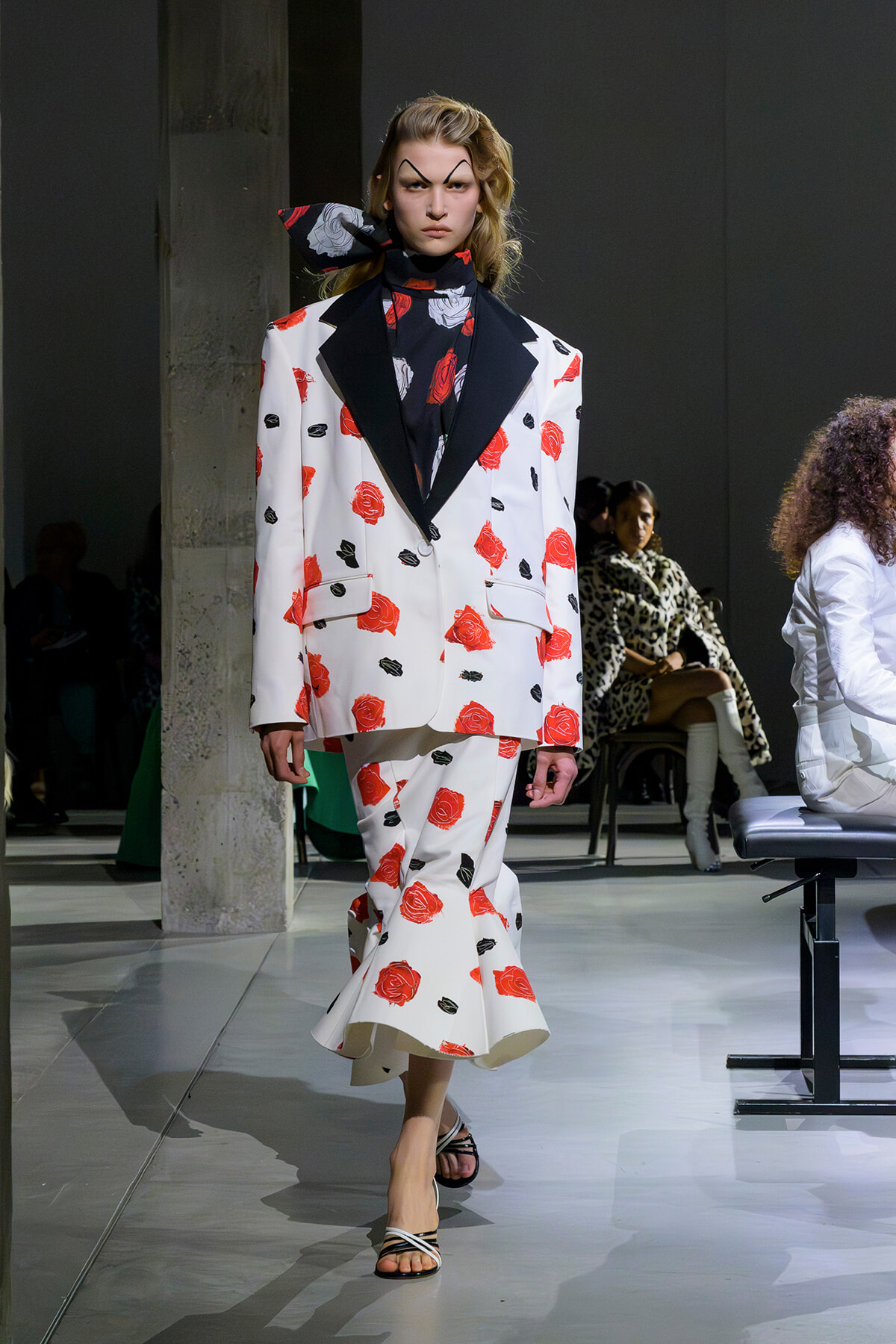 Model on runway wearing a white rose-print suit with black lapels and a dark scarf, walking the catwalk.