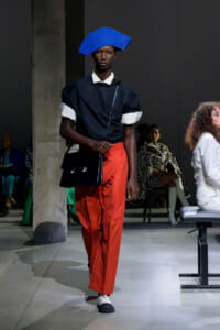 Model on runway wearing a bright blue angular hat, navy top with white cuffs, and orange wide-leg pants, carrying a black shoulder bag.