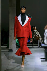 Model walking a runway in a red tailored suit with oversized white lapels, black top, and a flared mermaid-style bottom while wearing beige heels.