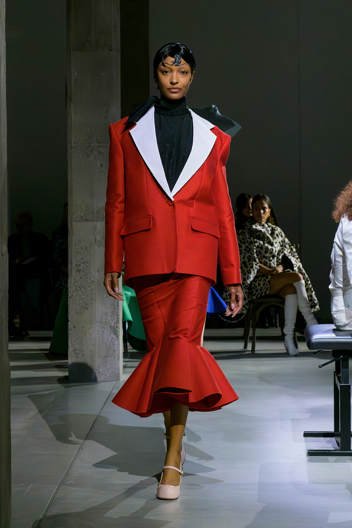 Model walking a runway in a red tailored suit with oversized white lapels, black top, and a flared mermaid-style bottom while wearing beige heels.