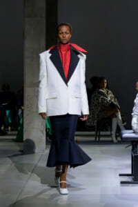 Model on a runway wearing a white oversized blazer with black peaked lapels, red turtleneck, and navy mermaid skirt, white heels.