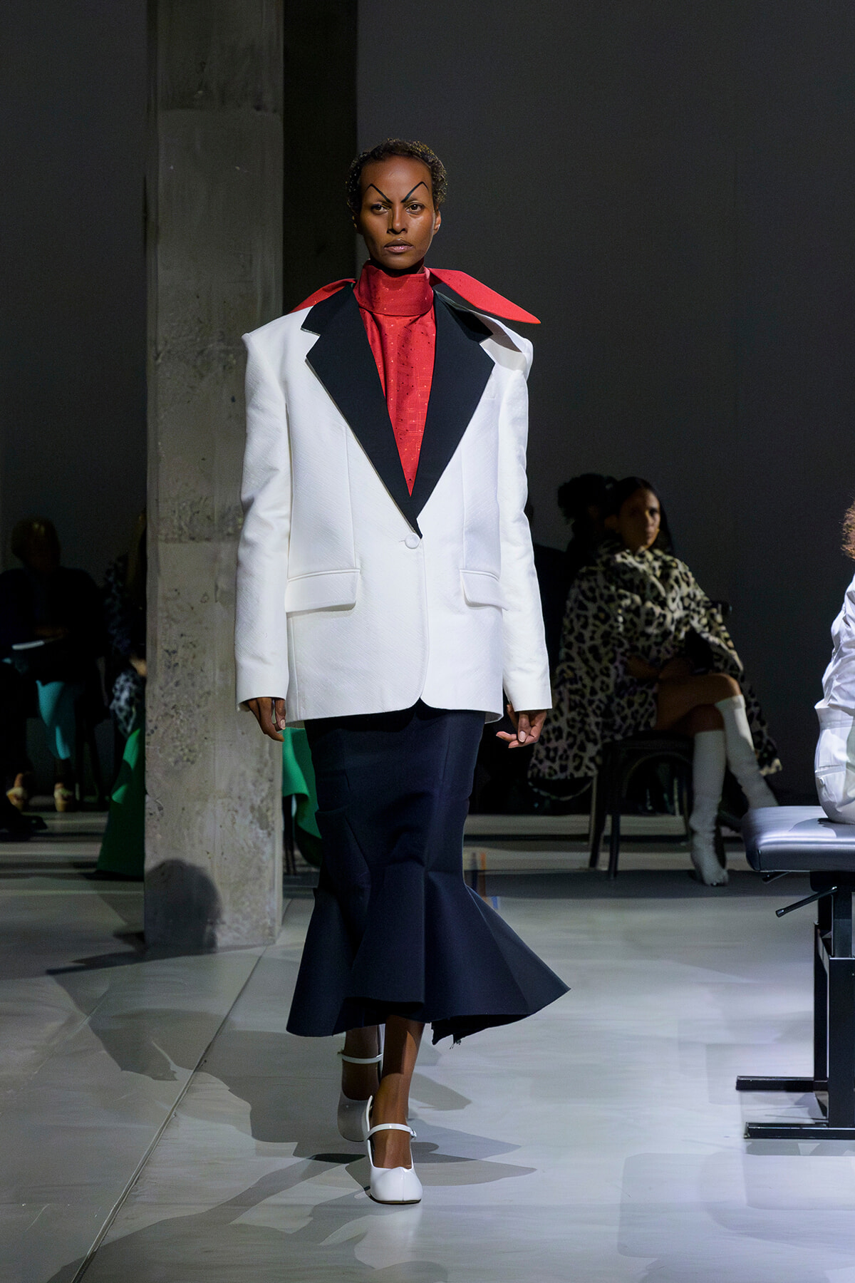 Model on a runway wearing a white oversized blazer with black peaked lapels, red turtleneck, and navy mermaid skirt, white heels.