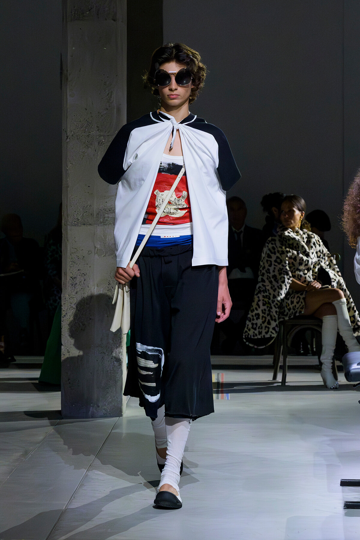 Model walking a fashion runway in a white cape top with black sleeves, graphic red-and-white shirt, wide black pants, white leg wraps, and black flats, wearing round sunglasses