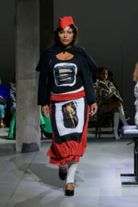 Model walking a runway in a black top with a white framed graphic, a red layered skirt, white leg wraps, black shoes, and a red origami-style headpiece.