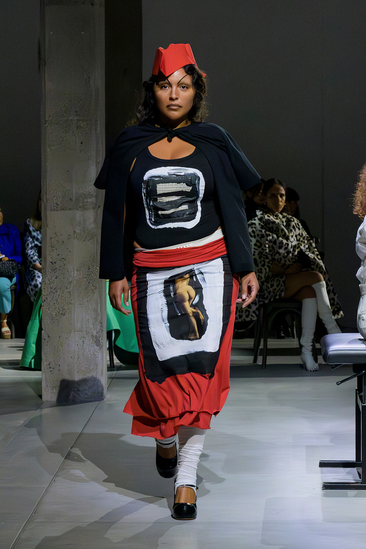Model walking a runway in a black top with a white framed graphic, a red layered skirt, white leg wraps, black shoes, and a red origami-style headpiece.