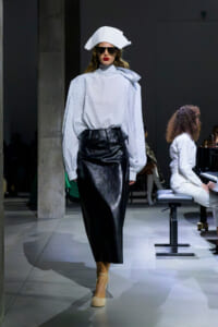 Model on a runway wearing a white oversized blouse with puff sleeves and a high neck, a black leather midi skirt, beige heels, and a white sculpted hat with dark sunglasses.