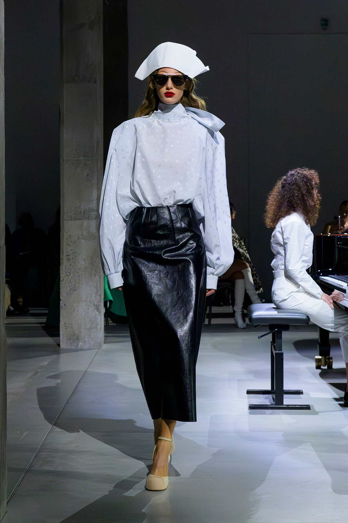 Model on a runway wearing a white oversized blouse with puff sleeves and a high neck, a black leather midi skirt, beige heels, and a white sculpted hat with dark sunglasses.
