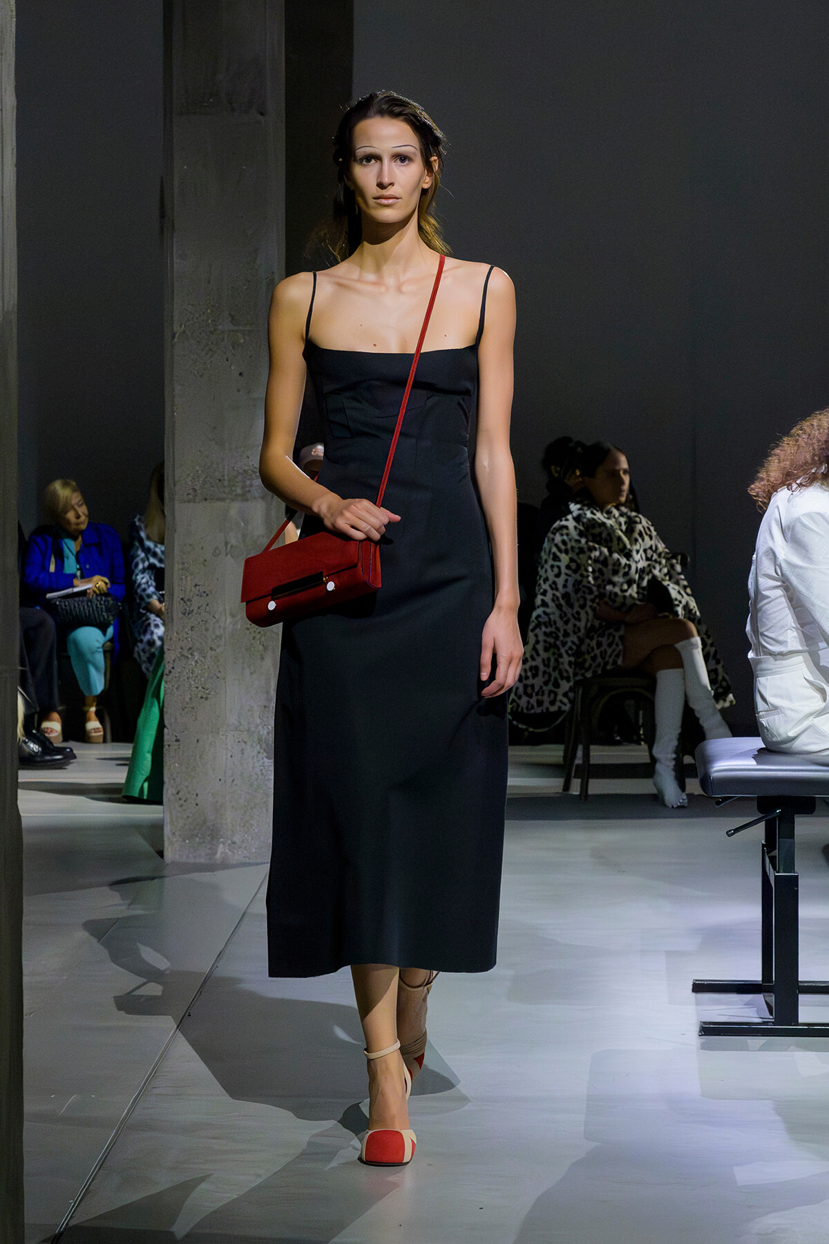 Model walking a runway in a black slip dress with a red crossbody bag and two-tone red-and-cream heels.
