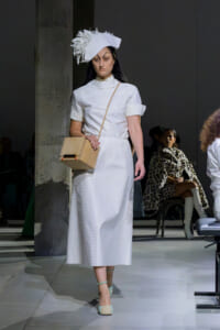 Female model walks a runway in a white midi dress with a sculptural white hat, beige crossbody bag, and mint heels.