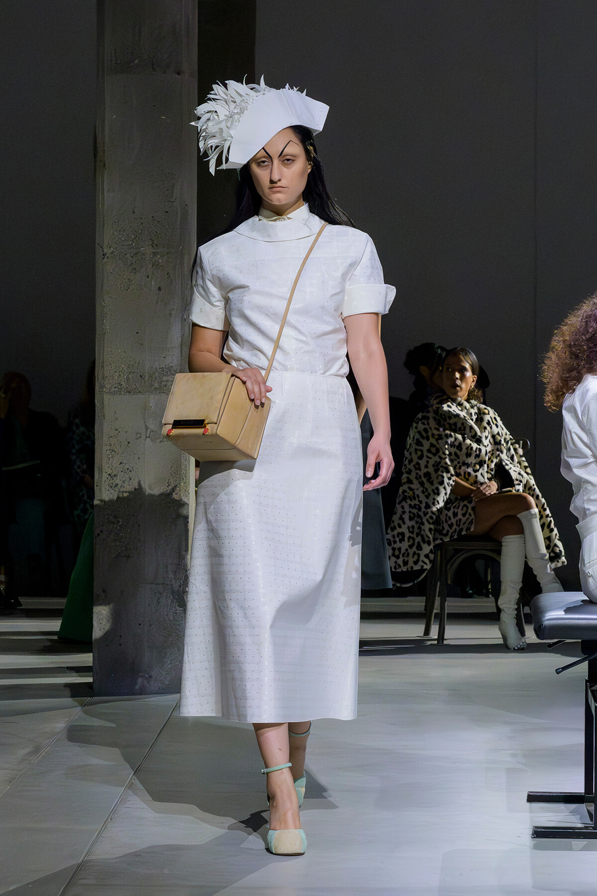Female model walks a runway in a white midi dress with a sculptural white hat, beige crossbody bag, and mint heels.