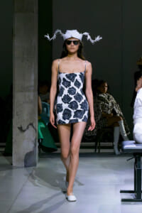Model walking the runway in a white mini dress with a navy rose print, a white feathered hat, sunglasses, and white flats.