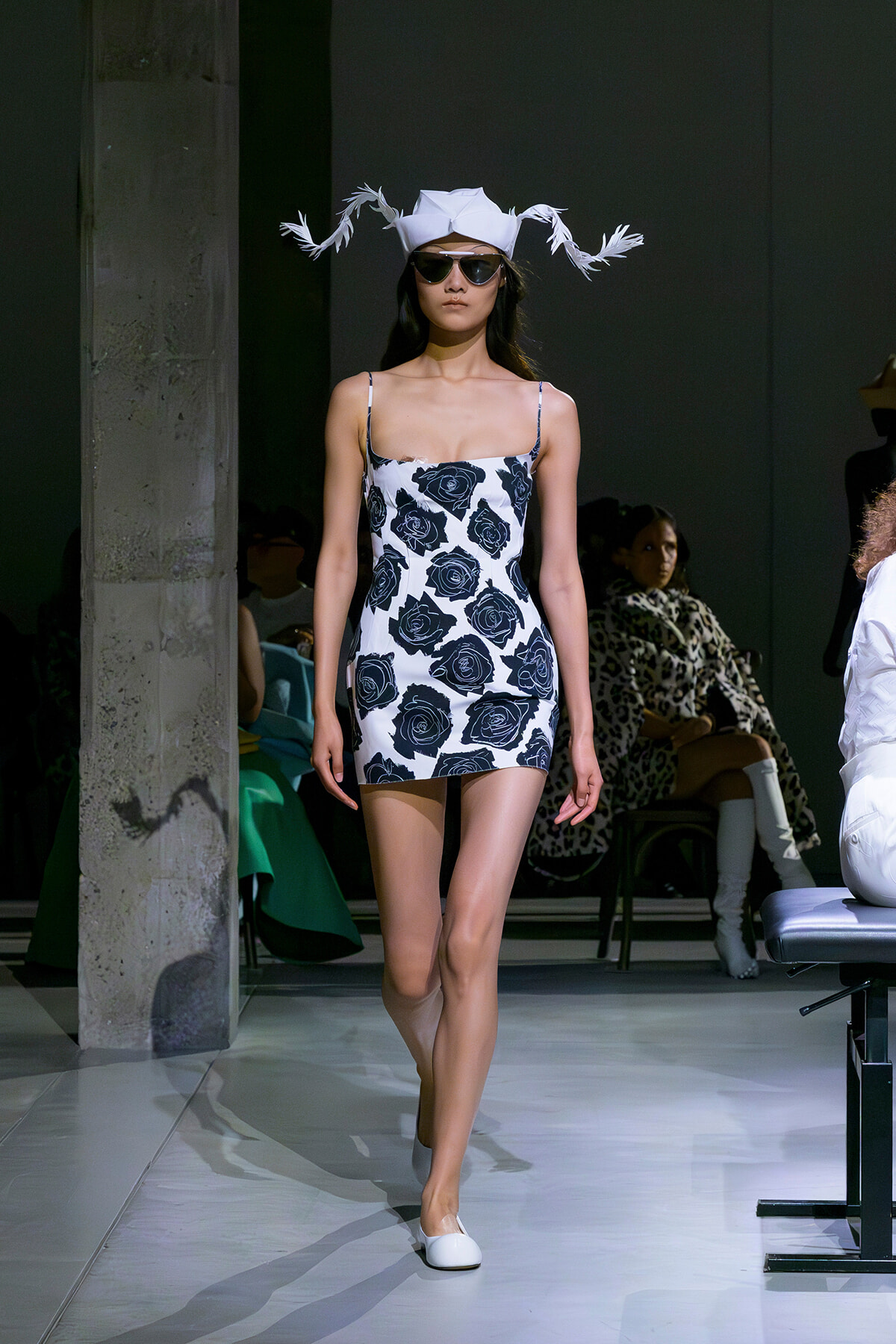 Model walking the runway in a white mini dress with a navy rose print, a white feathered hat, sunglasses, and white flats.
