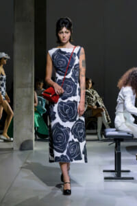 Model walks the runway in a sleeveless white dress with large black rose prints and a red crossbody bag.