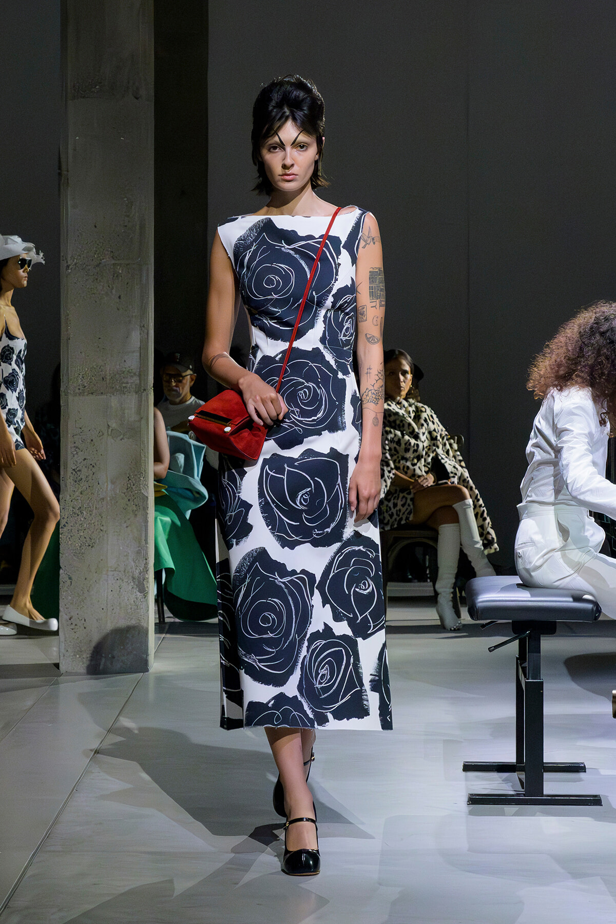 Model walks the runway in a sleeveless white dress with large black rose prints and a red crossbody bag.