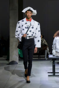 Model walks a runway in a white rose-pattern jacket with a red collar, paired with black pants, black flats, a white sculptural hat, and a black shoulder bag.