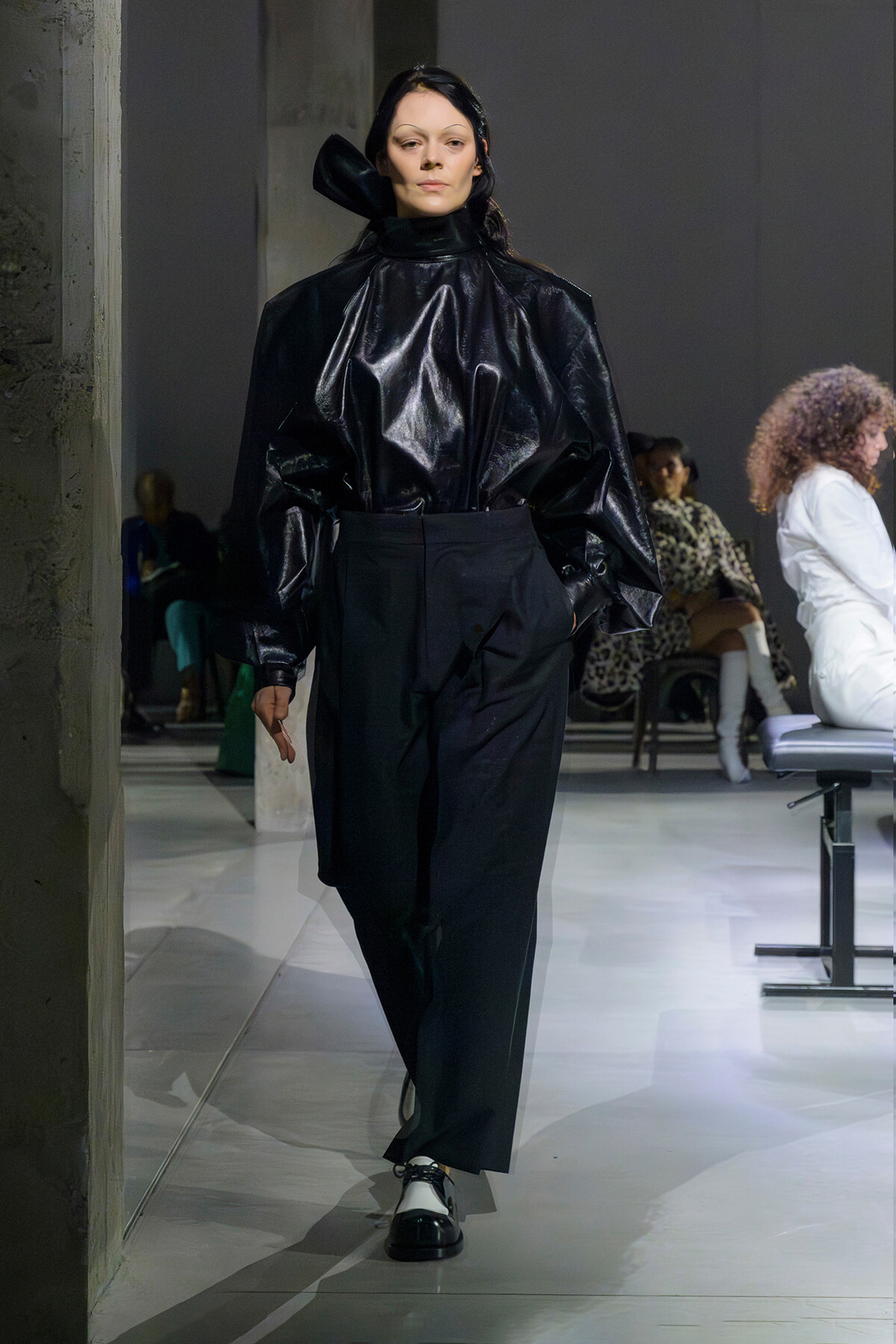 Model walks the runway in a shiny black high-neck top with exaggerated puff sleeves and high-waisted black trousers, black-and-white oxford shoes visible.