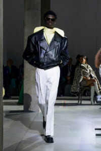 Model walking a runway in an oversized black leather double-breasted jacket, pale yellow scarf, white pants, and dark sunglasses.