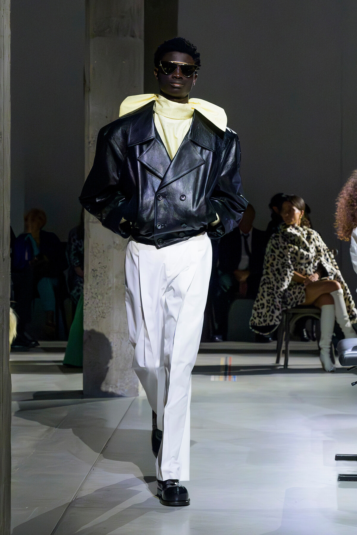 Model walking a runway in an oversized black leather double-breasted jacket, pale yellow scarf, white pants, and dark sunglasses.