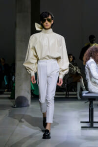 Model on a runway wearing a cream voluminous top with puff sleeves and white high-waisted pants, black loafers, and aviator sunglasses.