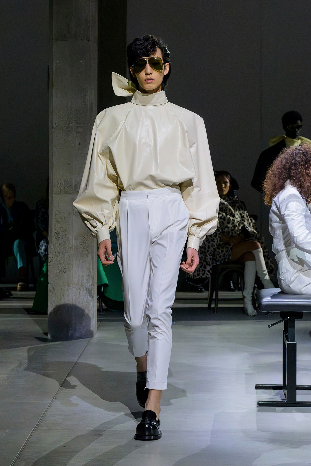 Model on a runway wearing a cream voluminous top with puff sleeves and white high-waisted pants, black loafers, and aviator sunglasses.