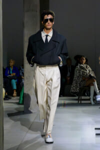 Runway model in an oversized dark navy blazer, white shirt and tie, cream wide trousers, and sunglasses walking the catwalk.