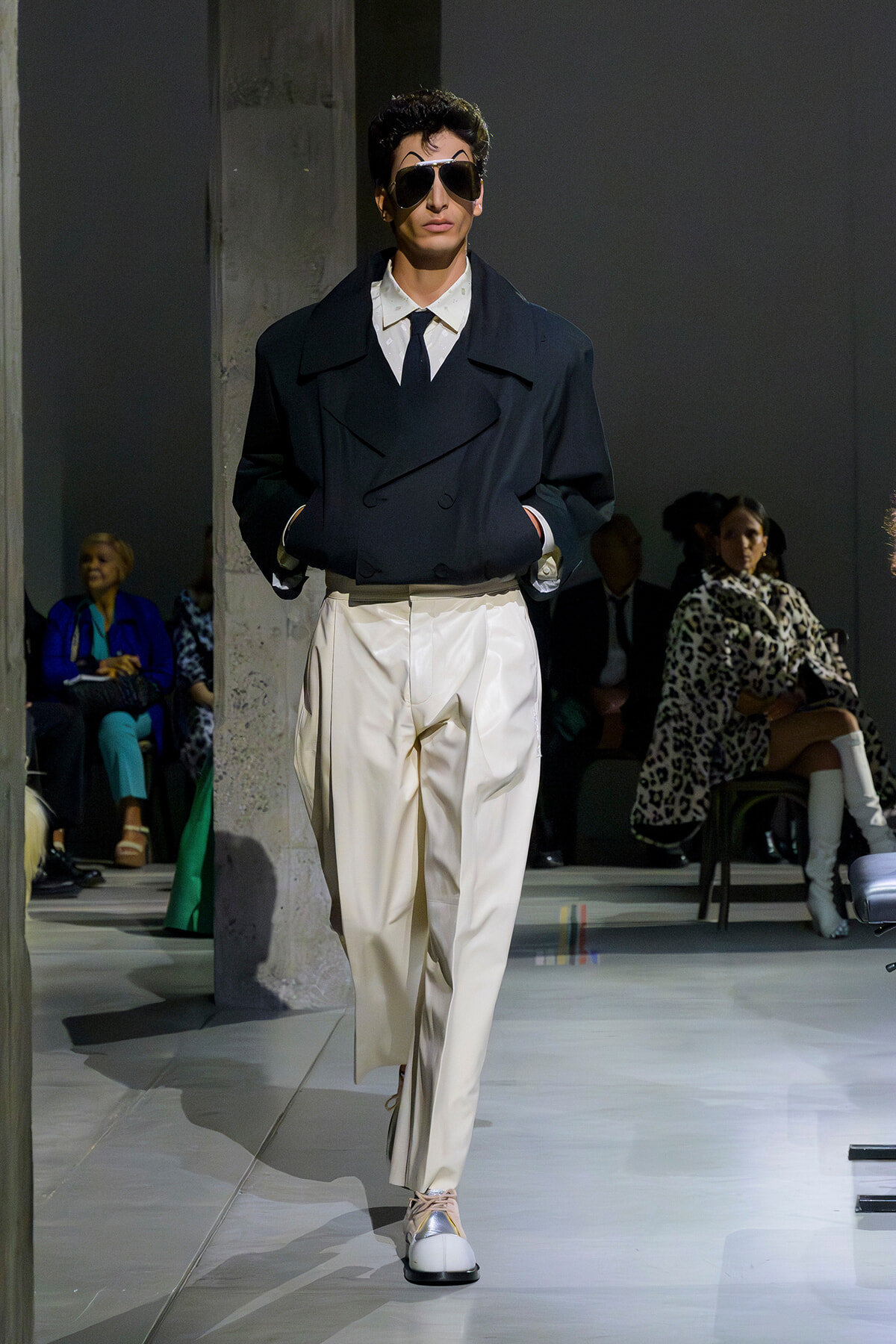 Runway model in an oversized dark navy blazer, white shirt and tie, cream wide trousers, and sunglasses walking the catwalk.