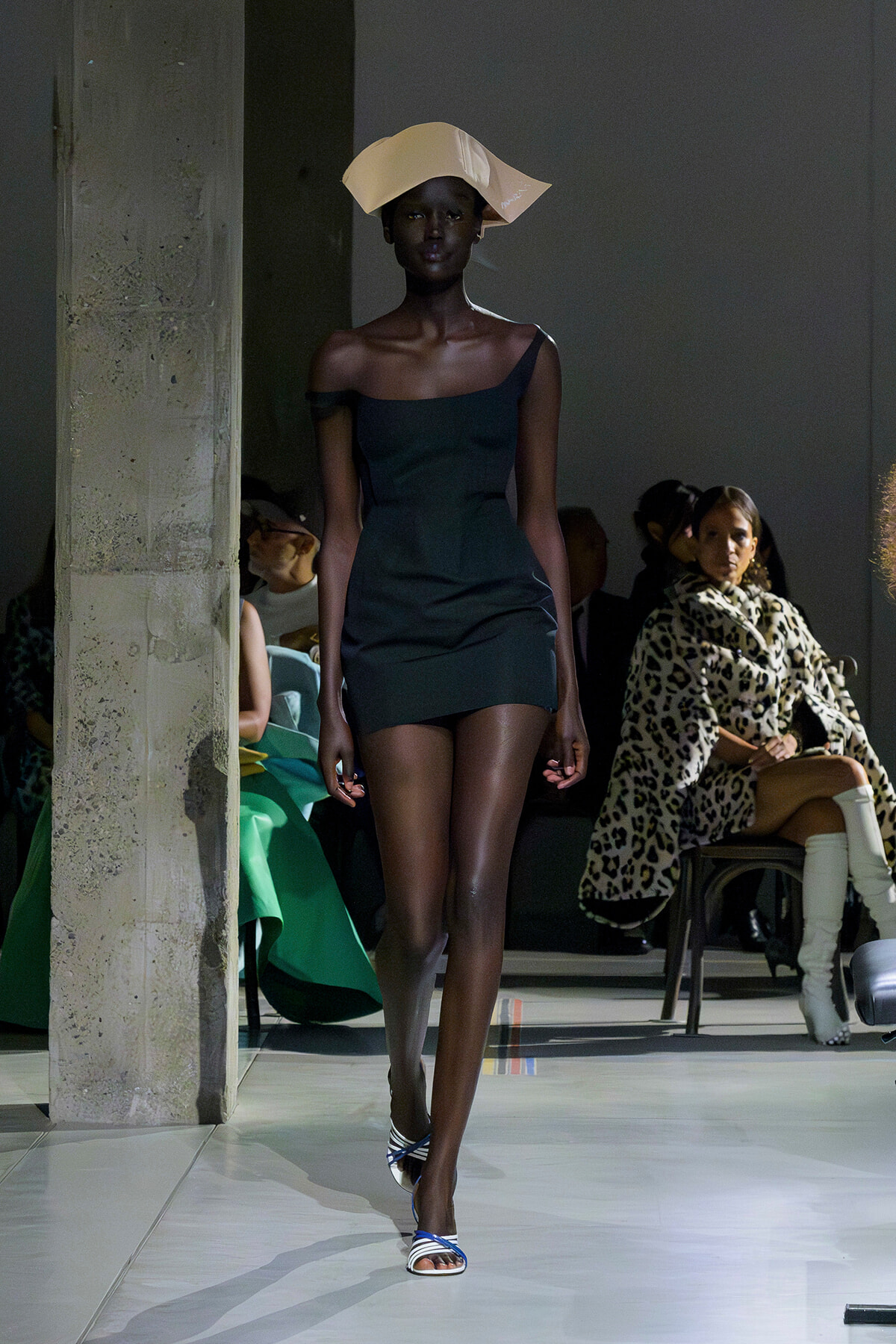 Model walks the runway in a dark one-shoulder mini dress, beige sculptural hat, and blue-white striped heeled sandals; audience seated in the background.