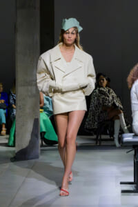 Model walking the runway in an oversized cream trench with a sculpted mint hat and pale pink sandals.