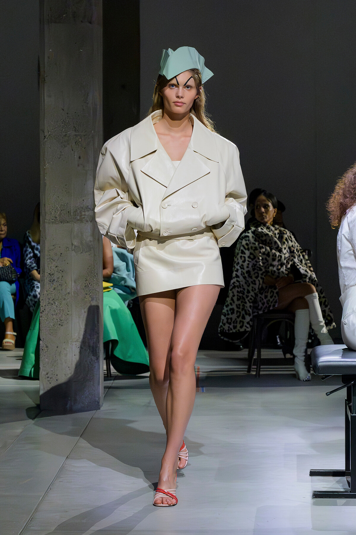 Model walking the runway in an oversized cream trench with a sculpted mint hat and pale pink sandals.