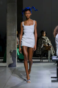 Model walking a fashion show runway in a white mini dress with a blue feathered headpiece and strappy sandals, audience in background.