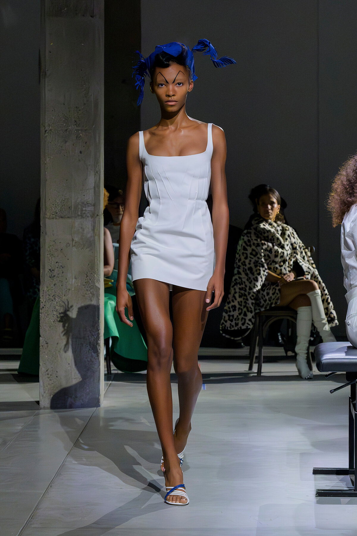Model walking a fashion show runway in a white mini dress with a blue feathered headpiece and strappy sandals, audience in background.