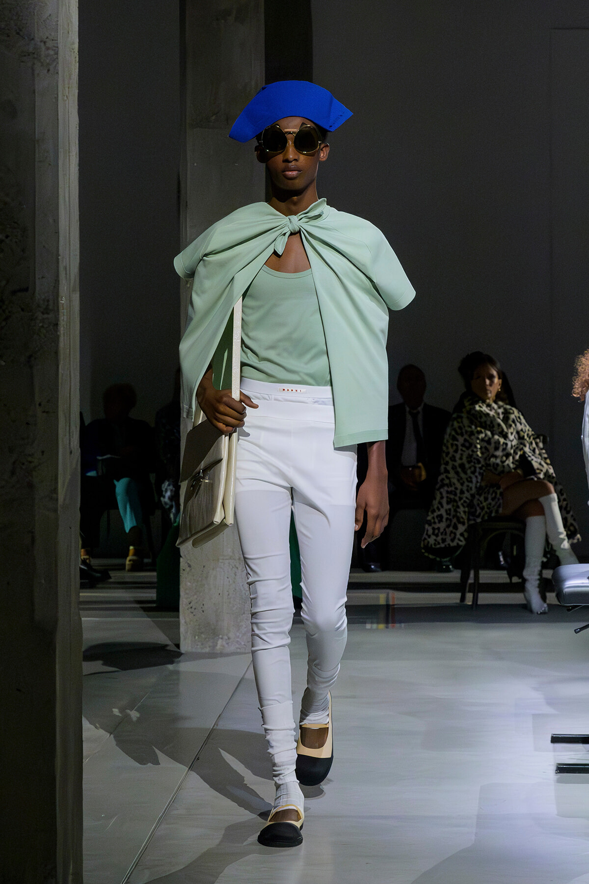 Model on a runway wearing a pastel mint cape tied at the neck over a mint top, white pants, oversized blue hat, sunglasses, and a beige handbag.