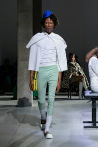 Model walking a fashion runway in a white cape top, mint pants, white socks, and a blue headpiece, carrying a yellow handbag.