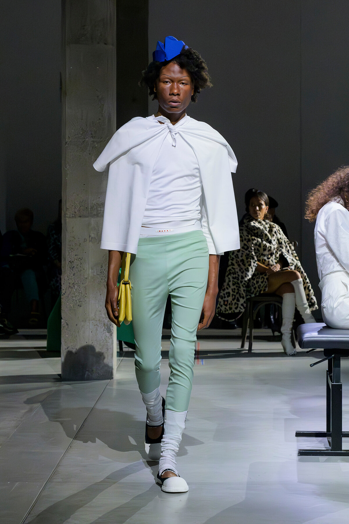Model walking a fashion runway in a white cape top, mint pants, white socks, and a blue headpiece, carrying a yellow handbag.