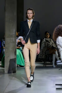 Model walking a fashion runway in a black blazer, beige pants, white cuffs, and a small black crossbody bag.
