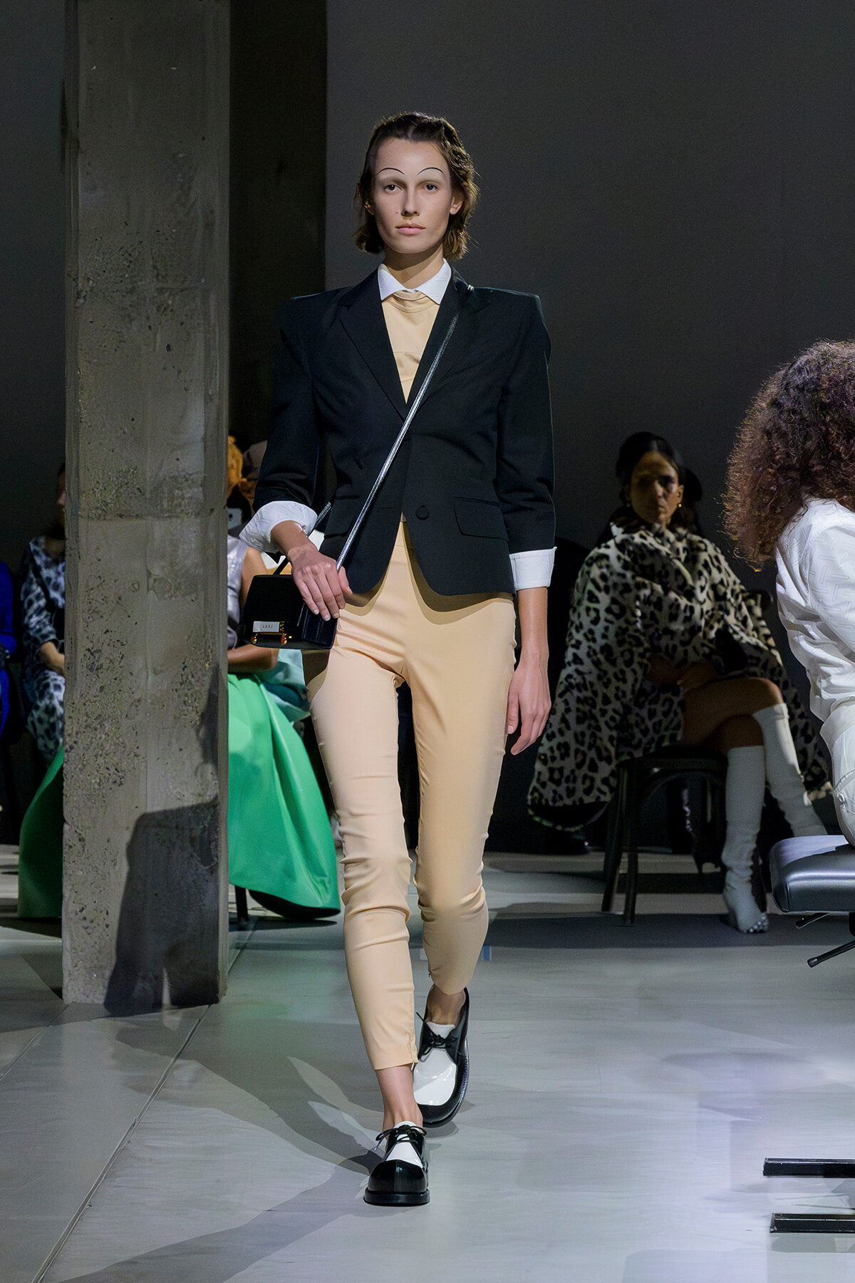 Model walking a fashion runway in a black blazer, beige pants, white cuffs, and a small black crossbody bag.
