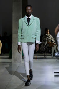 Model walks a runway in a mint green blazer, white pants, and black shoes, showcasing a modern, minimalist look.