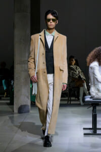 Model on a runway wearing a beige long coat over a black vest and white pants, with sunglasses and a light gray bag over the shoulder.