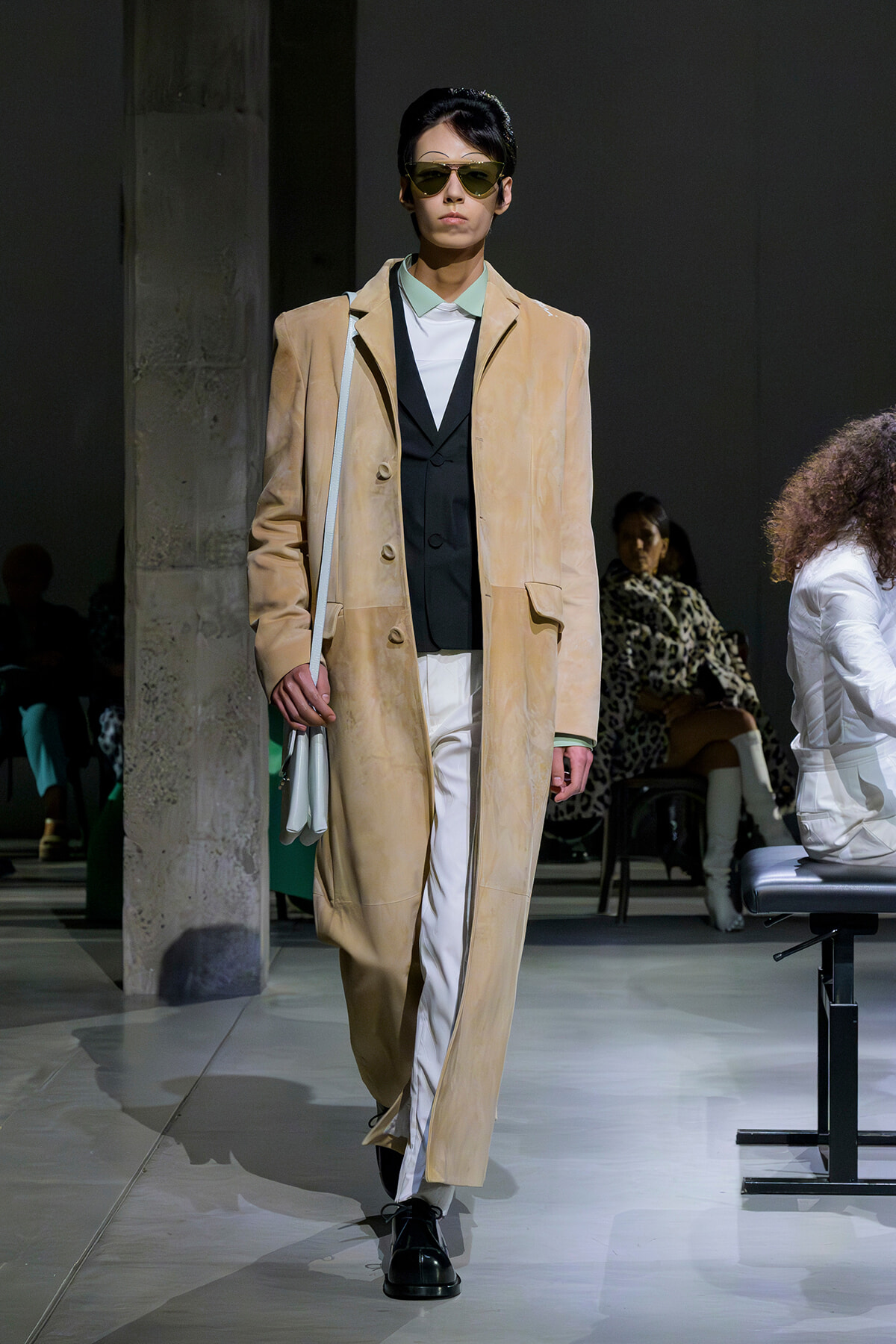 Model on a runway wearing a beige long coat over a black vest and white pants, with sunglasses and a light gray bag over the shoulder.