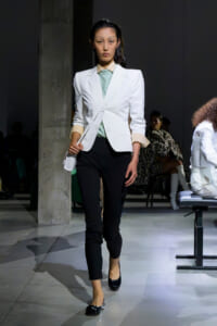 Female model walking a runway in a white blazer, pale green blouse, black pants, and black flats, holding a small white bag in front of her.