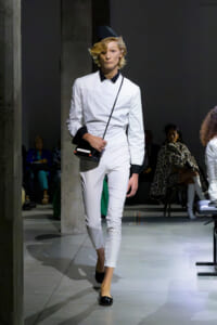 Model walks a fashion runway wearing a white tailored jumpsuit with black collar and cuffs, black flats, and a small black crossbody bag.