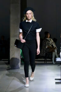 Model walking a runway in a black outfit with white cuffs and collar, beret, and crossbody bag.