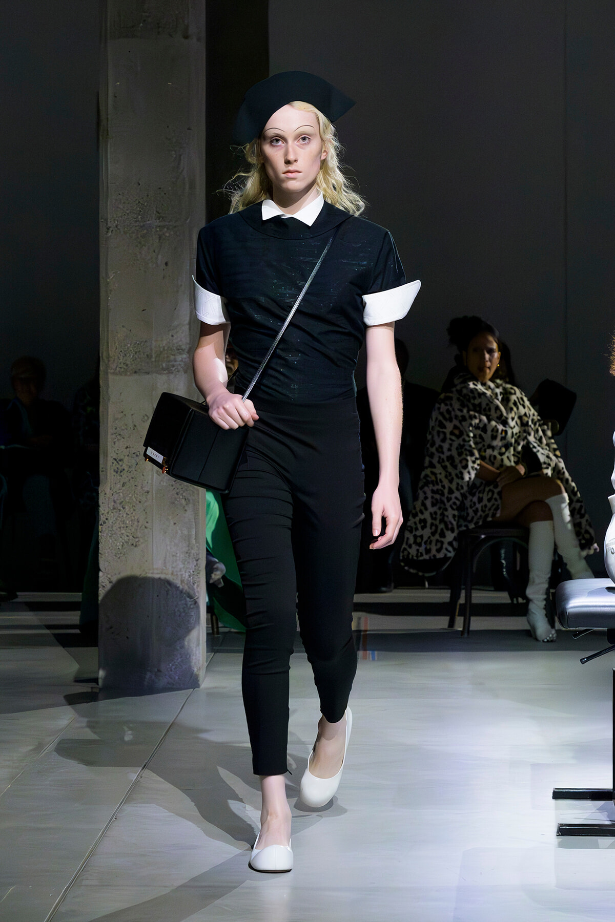 Model walking a runway in a black outfit with white cuffs and collar, beret, and crossbody bag.